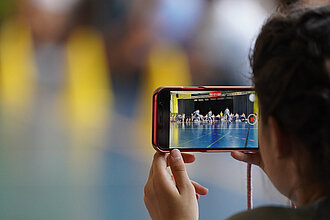 Ein Mädchen filmt mit der Handykamera einen Auftritt in einer Turnhalle.